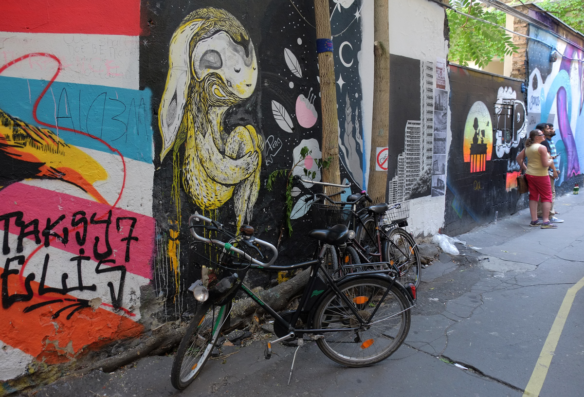 bike parked in front of a mural of a yellow human like creature with knees tucked up, people in the background looking at the street art 