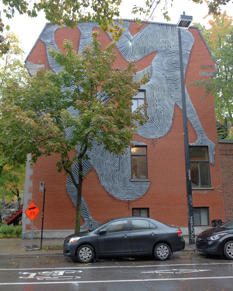 two large humanoid figures upside down, striped black and white, windows of the buildings are the faces of the creatures in the mural 