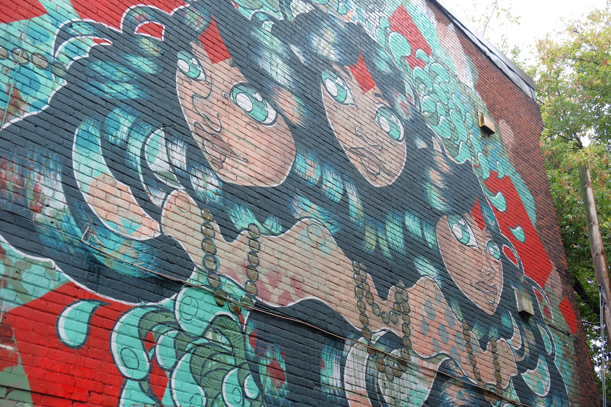 large mural on the side of the building, in greens on red background, three women with dark gren hair and bright green eyes