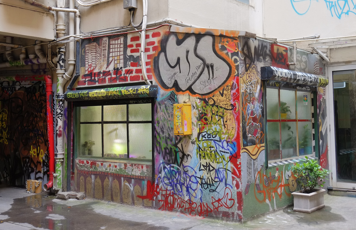 two windows in a small building in a lane, surrounding the windows is street art like a window box, awning, and then the wall space between the windows is decorated too 