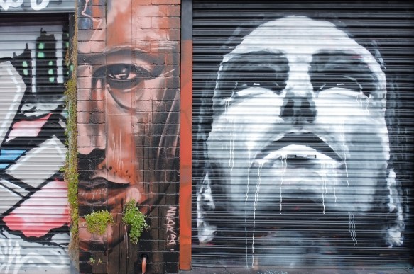two faces painted in Artists Lane in Windsor Melbourne, two male faces, one in black and white and the other in shades of brown. The black and white one is looking up, the brown one is only half a face (divided down the middle of the face, on eye, half a nose, and half a mouth. 