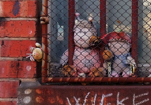 close up of three stuffies behind a rusty metal grille in front of a window. 