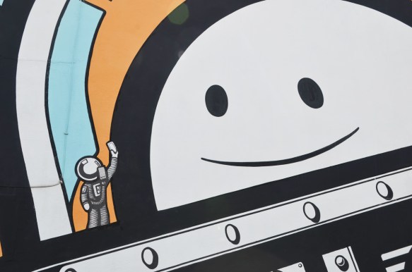 close up of big smiling astronaut face looking at a tiny astronaut, in a mural by The London Police 