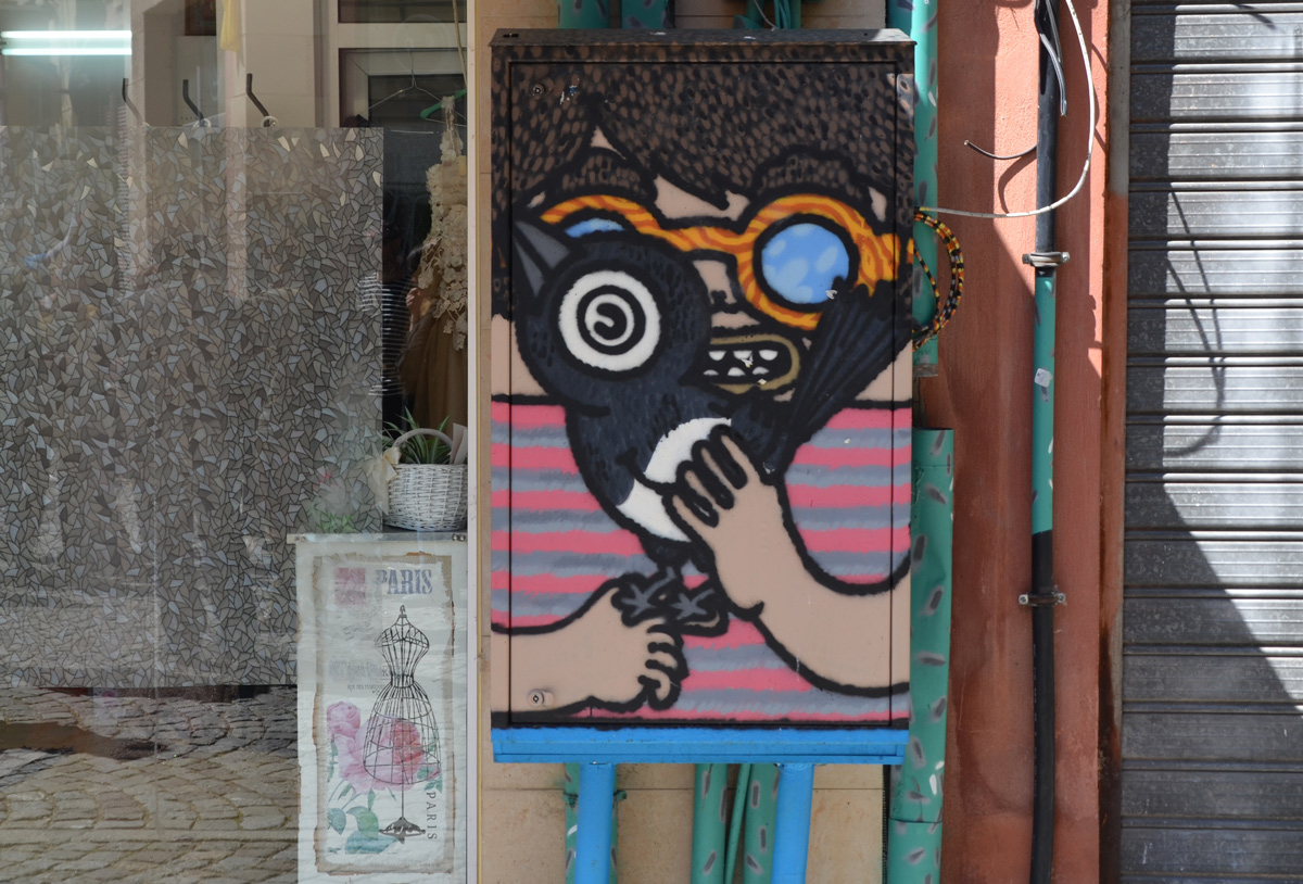 a painting on an electrical box on the street, of a boy with round orange glasses holding a large black bird 