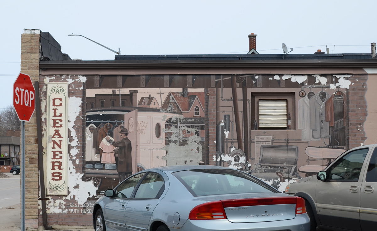 car parked in front of a mural showing the interior of an old cleaners with a man working inside hanging up a dress 