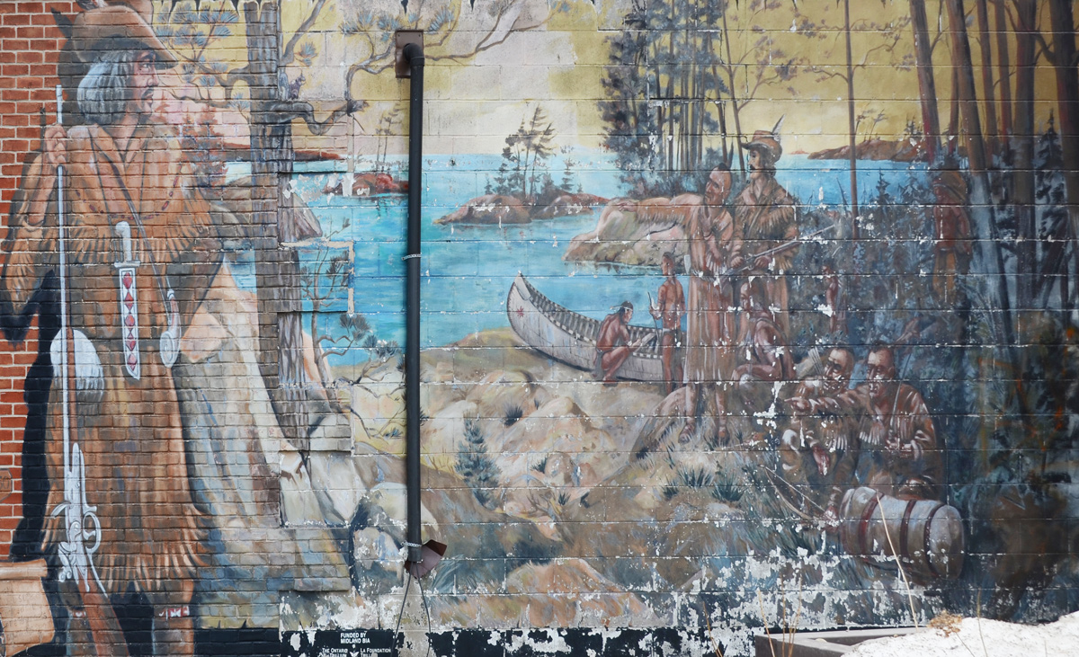 mural in bad shape (peeling paint) of a European explorer standing beside a lake scene, First Nations people and Europeans in canoes 