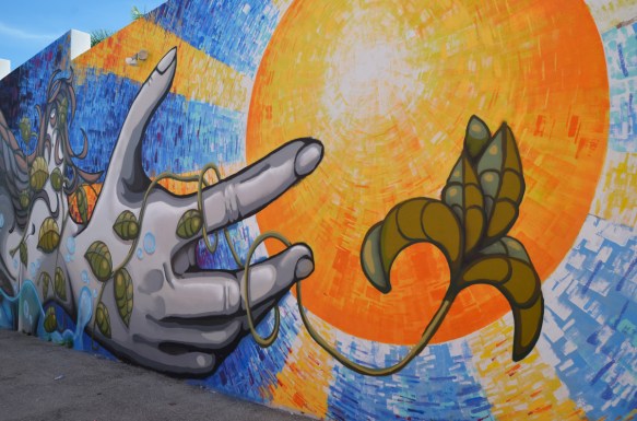 part of a mural by emo and remote, large yellow and orange sun in the middle, a hand is reaching for the sun, a vine is growing up the hand and fingers towards the sun 