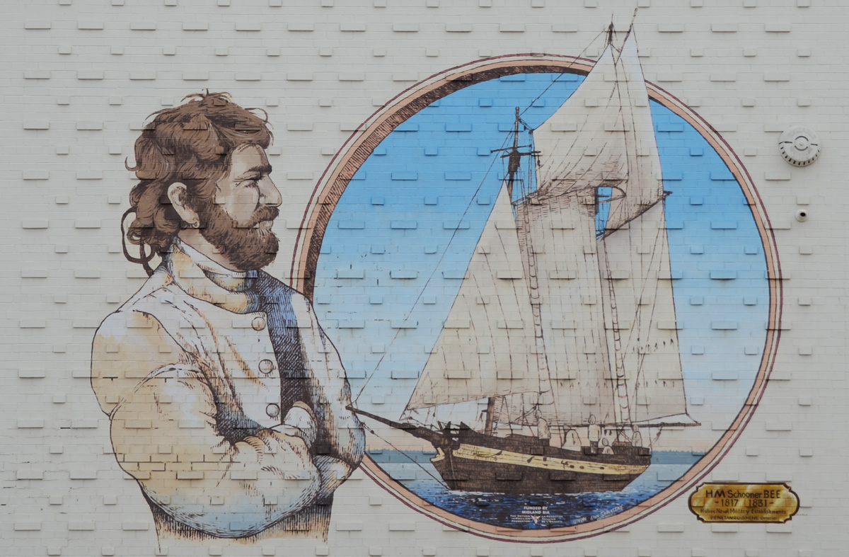 a man with a beard stands with his arms crossed looking at a picture of a schooner with 3 sails, the HMS Bee 