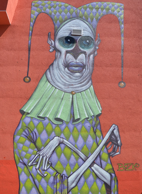 a large mural of an older man in jester outfit, with arms crossed in front of him, make up on his face 