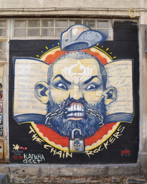 mural on a wall of the head ans shoulders of an older balding man with beard, moustache, and large white teeth. He has an angry look on his face. his collar is in Bulgarian colours, red, yellow and black. An open padlock is in his beard and a chain runs across the lower par tof his face but it lookslike he has just broken it with his teeth