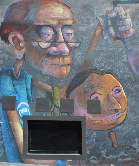 an older man with reading glasses and balding hair holds a wood puppet with a round head and a stick-like neck