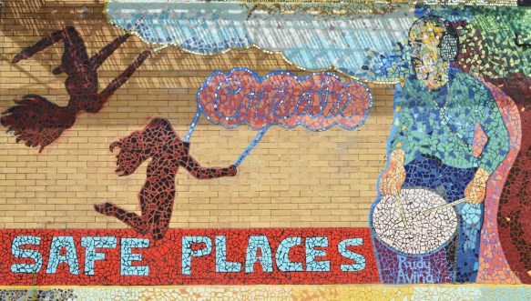 moasaic mural of tiles with words create safe places. A picture of a body outline on the ground with three red hearts, also shadows of two people with long hair on swings. A man plays a drum with the words Rudy Avina 