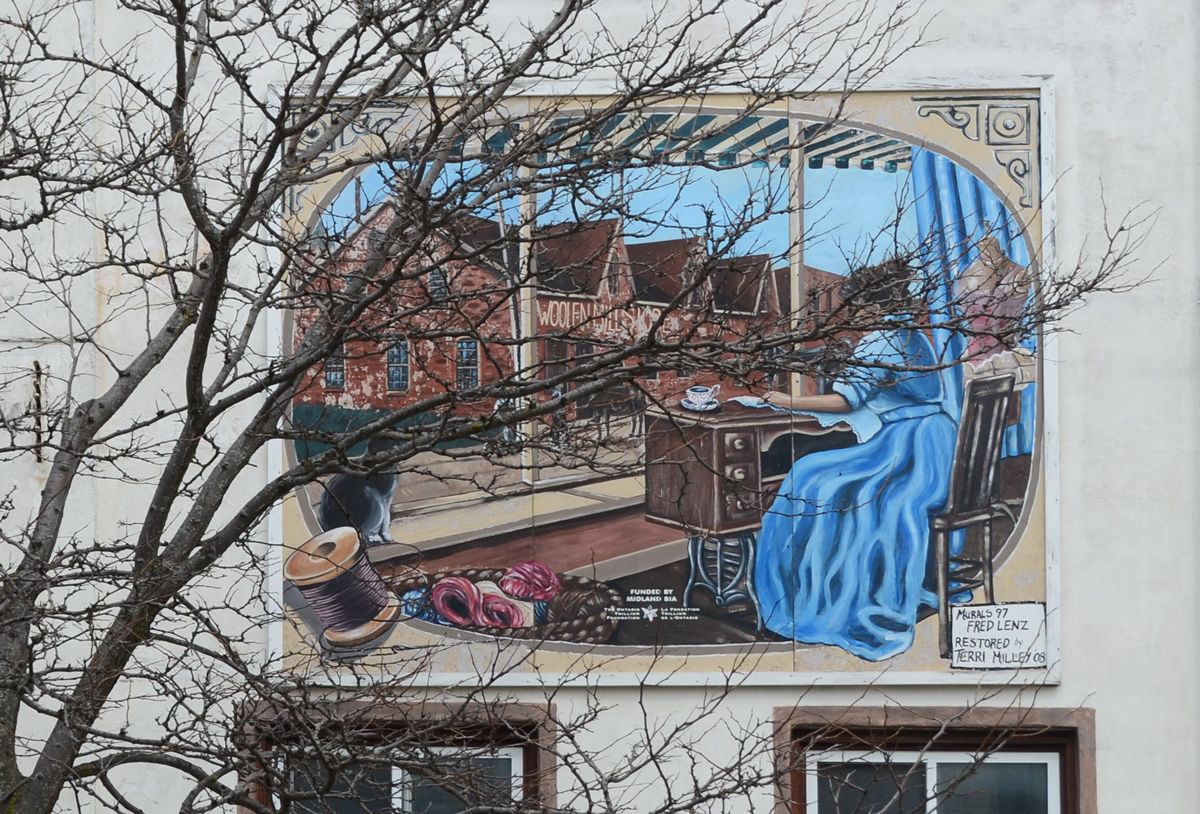 small mural behind a tree on the upper storey above a store, a woman in a long blue dress is sewing at on old fashioned sewing machine 