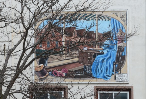 small mural behind a tree on the upper storey above a store, a woman in a long blue dress is sewing at on old fashioned sewing machine 