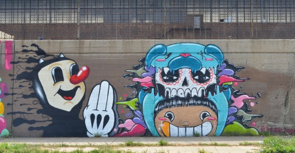 a mural with two creatures, a black cartoon cat head and shoulders, skinny arm with white gloved hand, long red nose. The other is a light blue, skull on top, smiley face with big teeth on the bottom