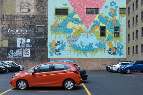 two murals on a wall on Wabash street in Chicago, on the wall Columbia college, beside a parking lot with a little bright orange car as well as other cars 