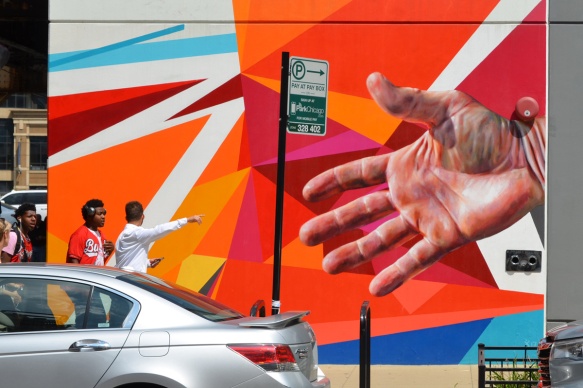 mural in bright oranges and reds, sharp edges, linear, with a realistic hand as well, people walking past it, one man with hand out pointing at something 