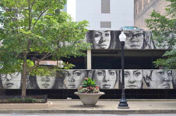 on the walls of a parking structure, a line of realistic faces in grey tones, two rows high