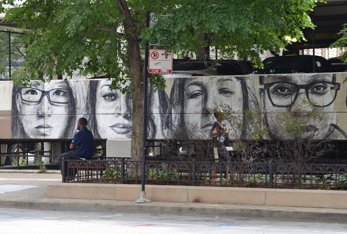 on the walls of a parking structure, a line of realistic faces in grey tones, a man sits on a bench beside it 