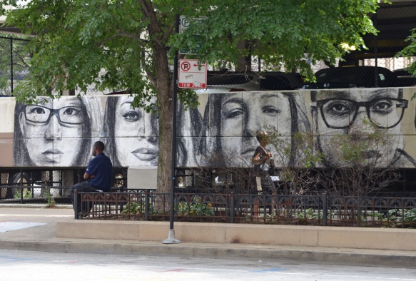 on the walls of a parking structure, a line of realistic faces in grey tones, a man sits on a bench beside it 