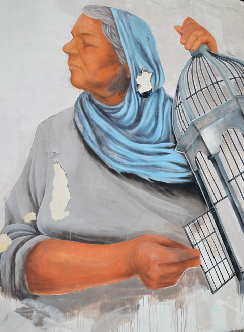 paint peeling on mural, woman in blue head scarf is holding open the door of a bird cage