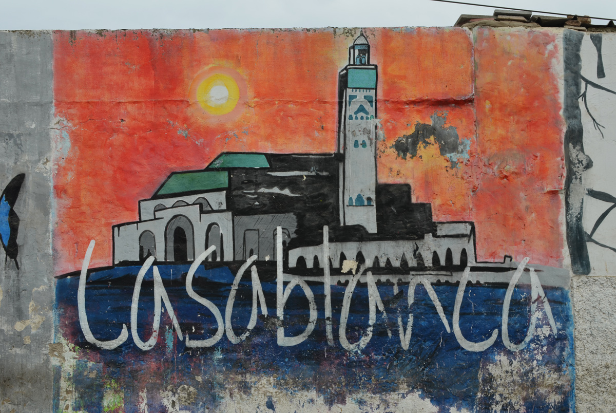 part of a series of murals on a Casablanca wall, the word Casablanca in white with a picture of the Hussan 2 mosque behind it, orange sky at sunset