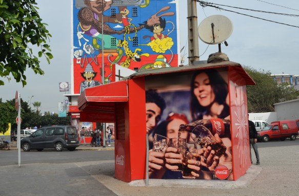 a kiosk covered with ads for coke products in front of a mural of cartoon characters by moka,