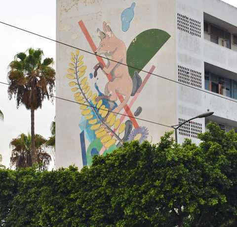 above the greenery of trees, a mural on the side of an apartment building, a foox, yellow leaves, and other pieces of nature 