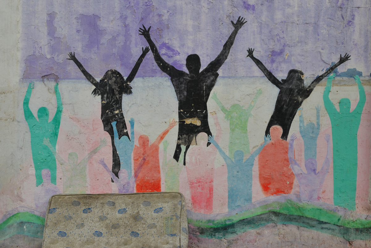 an old mattress sits in front of a mural of silhouette people shapes jumping, in black as well as red, green, and blue people