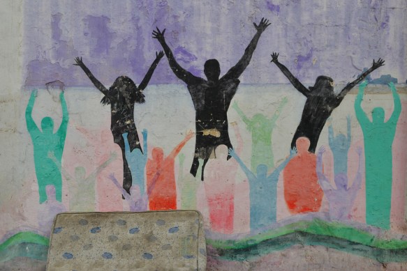 an old mattress sits in front of a mural of silhouette people shapes jumping, in black as well as red, green, and blue people