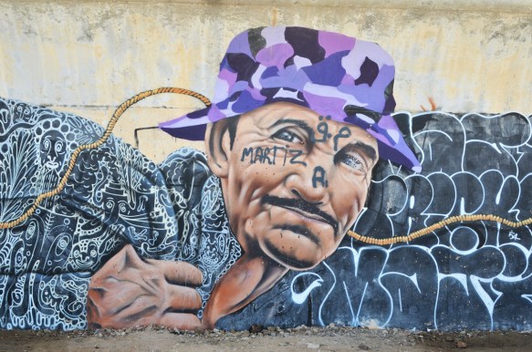 man's head, mural, wearing a purple camo hat and a black top with lots of white squiggles on it. A rope winds its way through the image 
