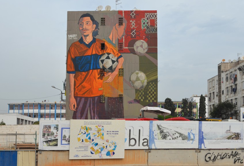 large mural by bakr of a young man in an orange blue T shirt holding a soccer ball or football, bottom part of mural is behind a fence