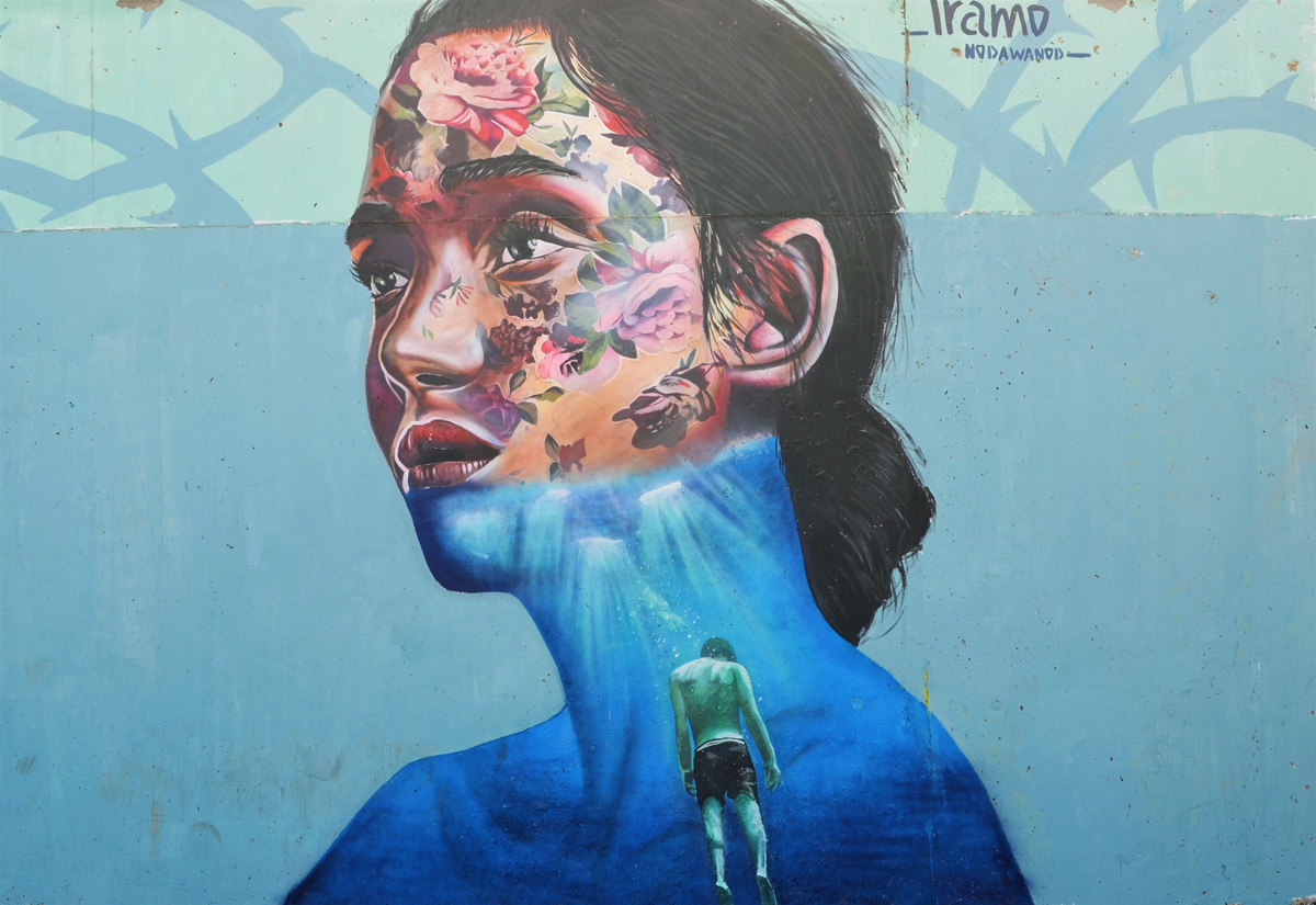 woman's head and shoulders, mural by Iramo Samir called Oxymoron, most of her face is covered with flowers, lower part of face and shoulders is blue water with a person who seems to be drowning