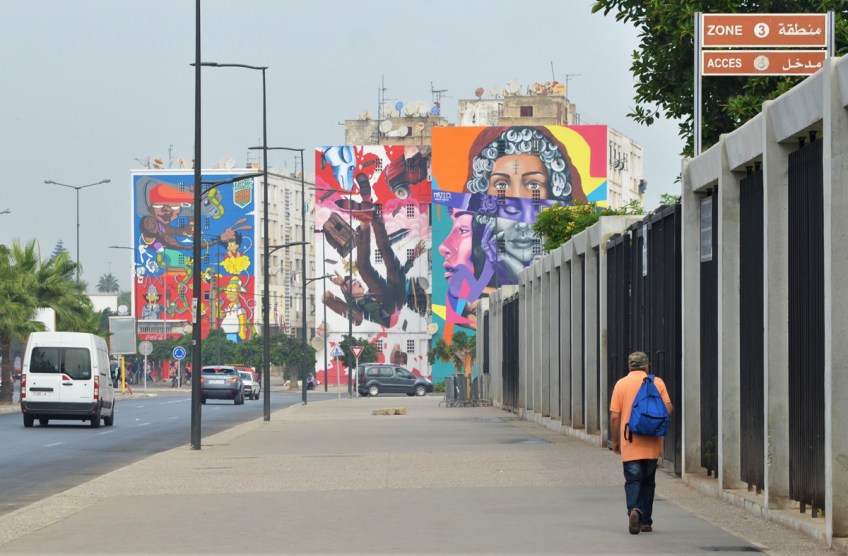 a man walks along a wide sidewalk, walking towards buildings with large colourful murals