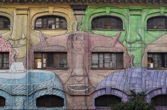 mural in Rome by Blu, on del porto fluviale, of large faces, where the eyes are windows in the buildings, brown face in middle