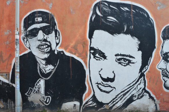 Elvis Presley and a rapper, large portraits by sten and lex, Italian street arts, in a mural on two sides of the street, of famous people and common people,