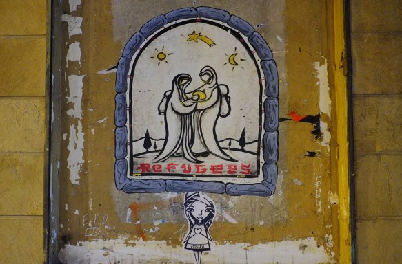 street art paste up of a nativity scene, Mary and Joseph standing together with Mary holding the baby Jesus, under night sky with moon and stars. The word refugees is written below the picture. All contained in a grey arch shape 