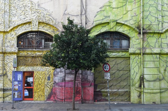 mural in Rome by Blu, on del porto fluviale, of large faces, where the eyes are windows in the buildings, small tree growing in front, green face and yellow face made of bananas 