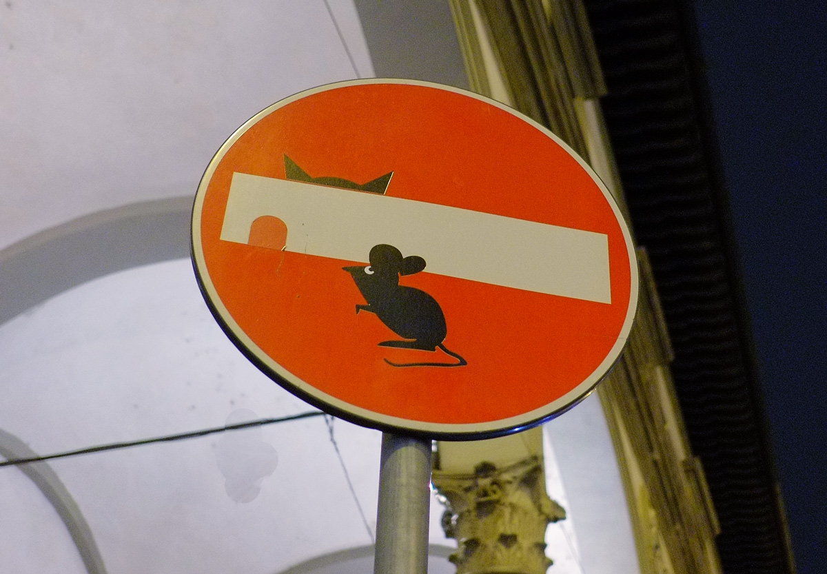 altered red and white no entry sign, graffiti by Clet Abraham, of a mouse beside a mouse hole, with a cat in the background