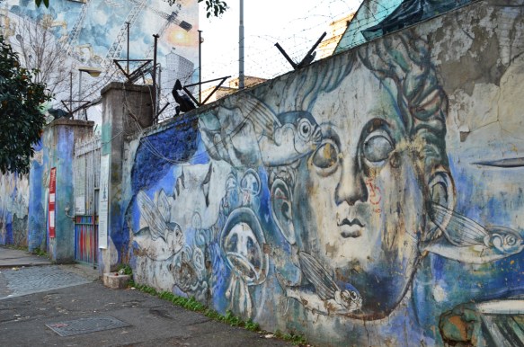 faces and fish on blue background, painted mural on wall with barbed wire above it, another mural on the wall beyond as well 