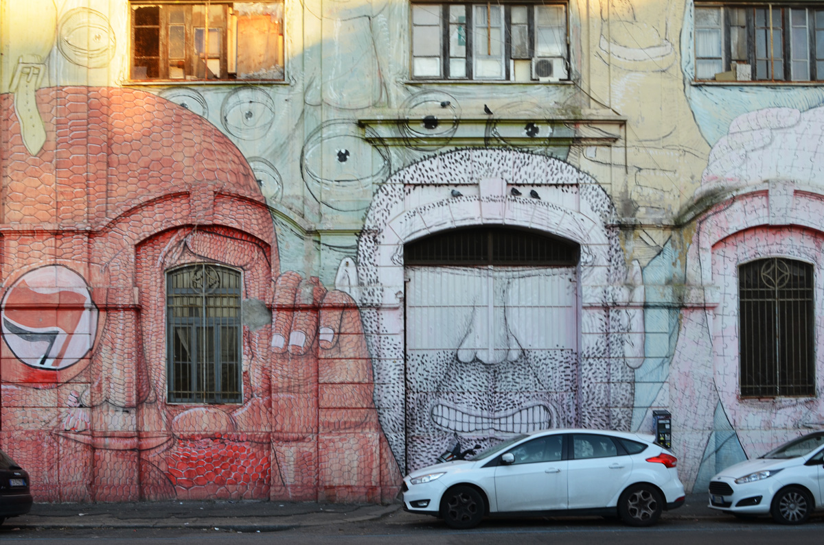 mural in Rome by Blu, on del porto fluviale, of large faces, where the eyes are windows in the buildings, red face and white face,