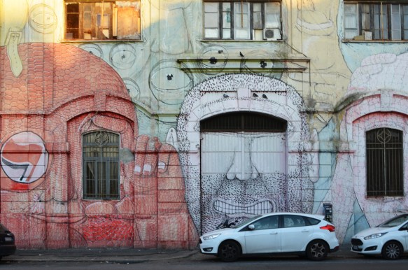 mural in Rome by Blu, on del porto fluviale, of large faces, where the eyes are windows in the buildings, red face and white face, 