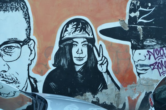 woman in hard hat giving peace symbol with hand, large portraits by sten and lex, Italian street arts, in a mural on two sides of the street, of famous people and common people,