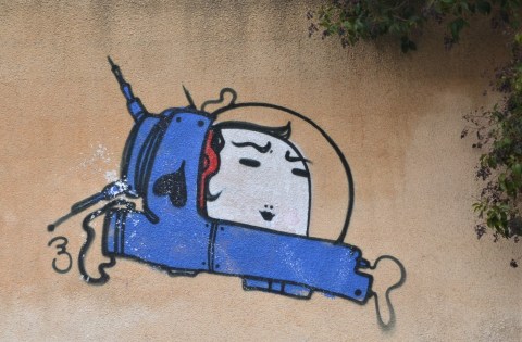 smallpainting on a concrete wall of a small blue spacecraft with a large white face inside it 