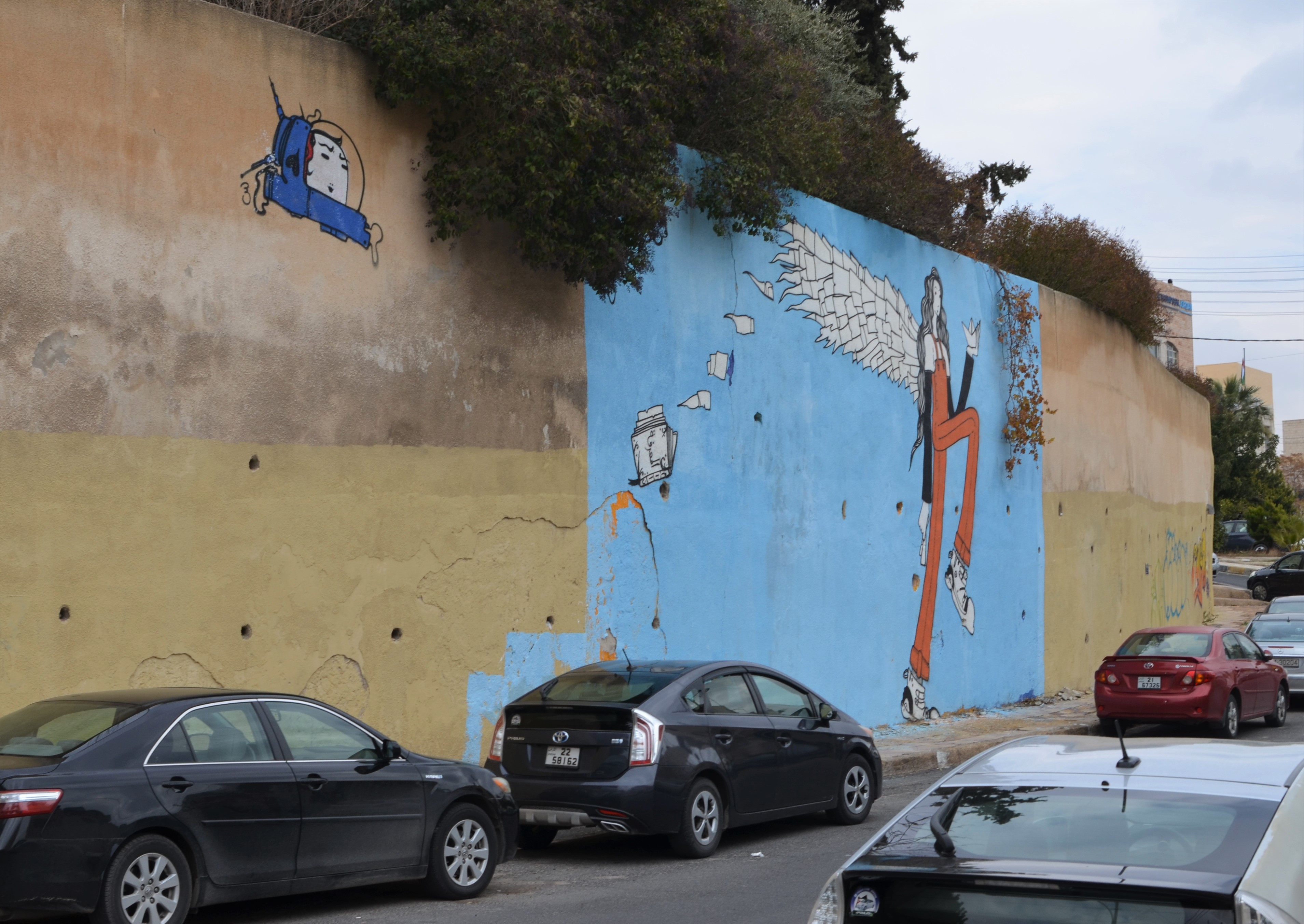 cars parked beside wall with blue mural of tall skinny woman with wings and small blue spaceship