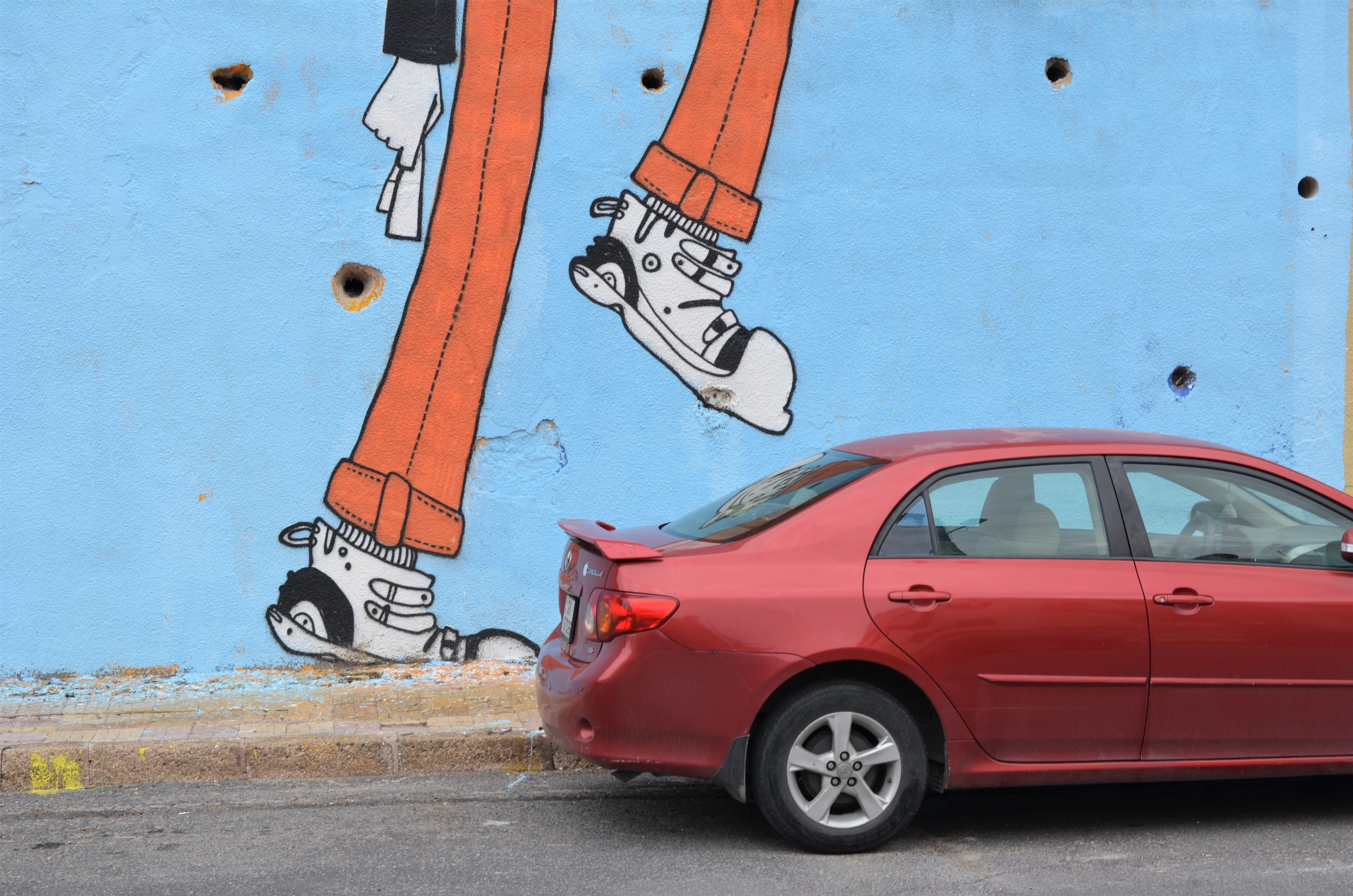 part of mural showing woman's legs, one leg raised looking like it's going to stomp on the red car that is parked beside the wall 