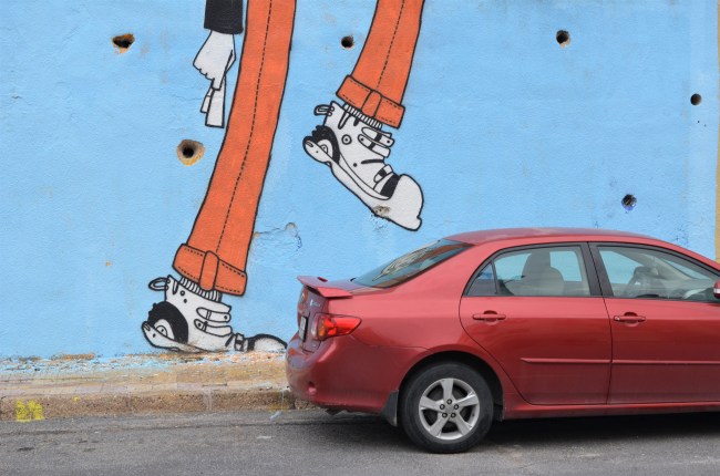 part of mural showing woman's legs, one leg raised looking like it's going to stomp on the red car that is parked beside the wall 