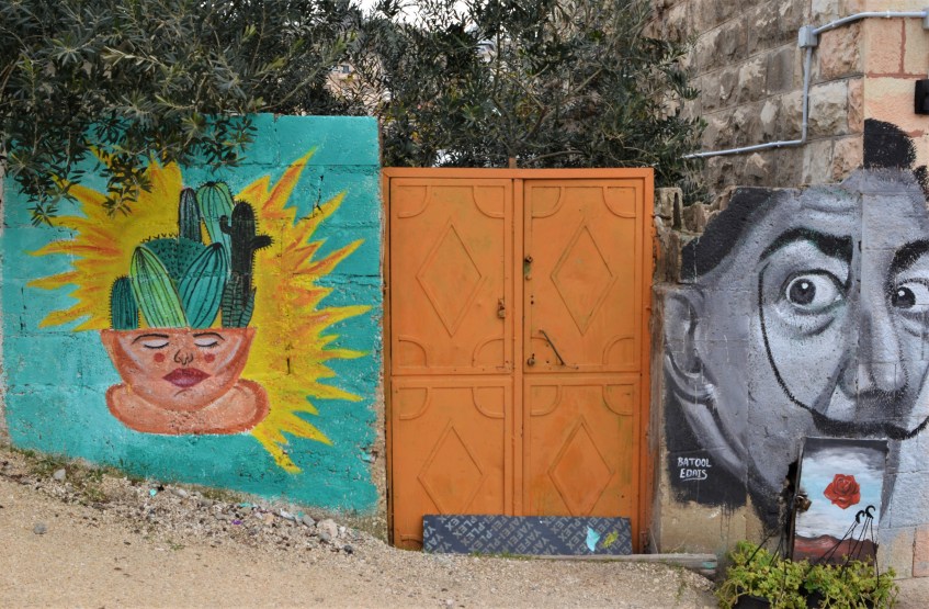 an orange metal gate in between two murals, on the left is face with a cactus instead of hair, and on the other side is a back and white picture of Salvator Dali
