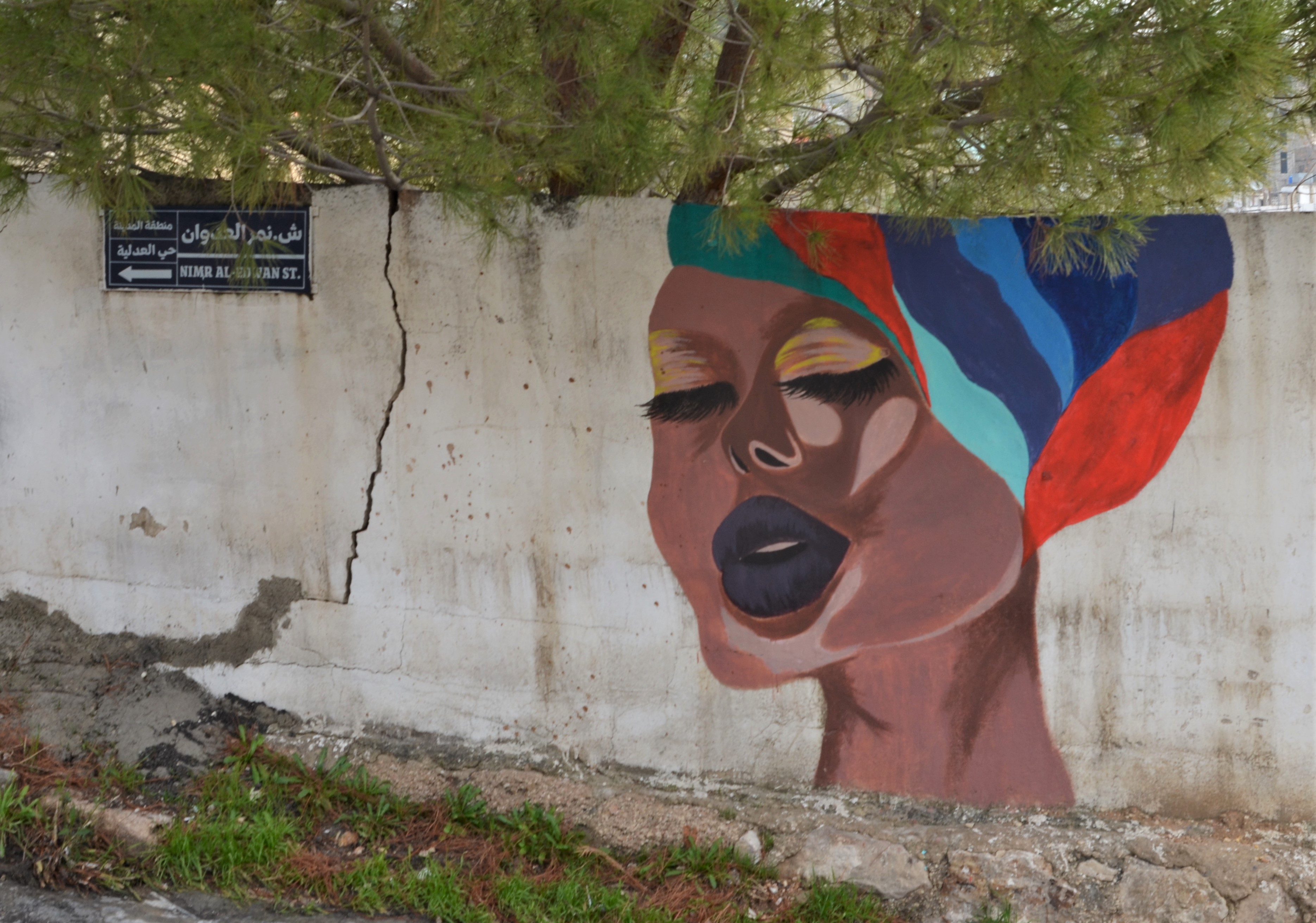 a woman in a colourful striped turban, singing with her eyes closed, mural pn a concrete wall, with green tree behind 
