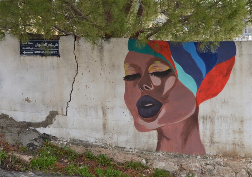 a woman in a colourful striped turban, singing with her eyes closed, mural pn a concrete wall, with green tree behind